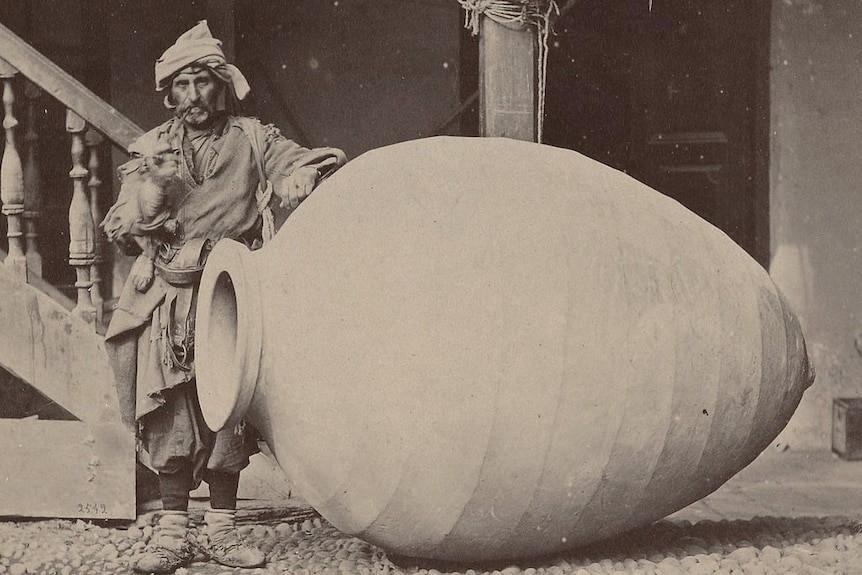 A black-and-white photo of a European tradesman in ragged clothes. He stands leaning on a giant clay jar.