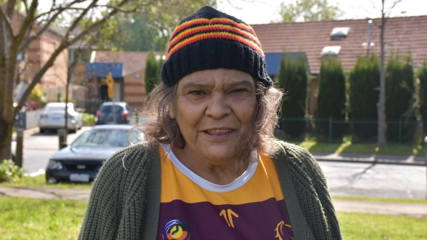A woman wearing a black, yellow and red beanie, and a dark green cardigan over a sports jumper stands on a suburban street.