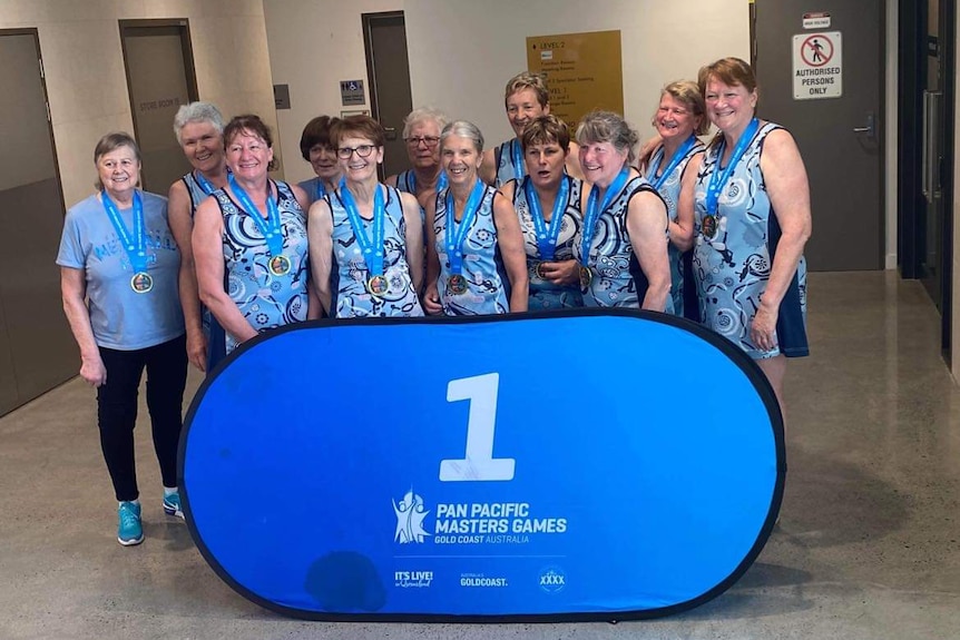 12 women over 60 wearing blue netball dresses stand behind a blue sign saying 1 pan pacific masters games