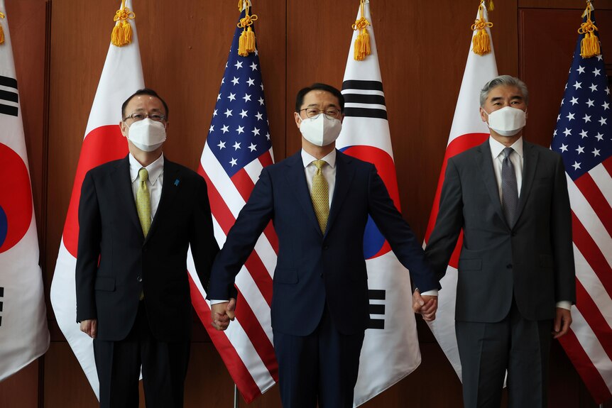 Takehiro Funakoshi, Kim Gunn and Sung Kim stand in front of Japanese, US and South Korean flags
