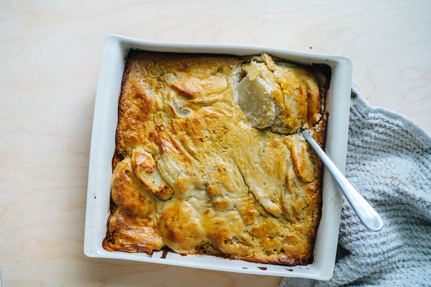 A spoon rests inside a baking dish filled with gratin, with a crispy brown top.