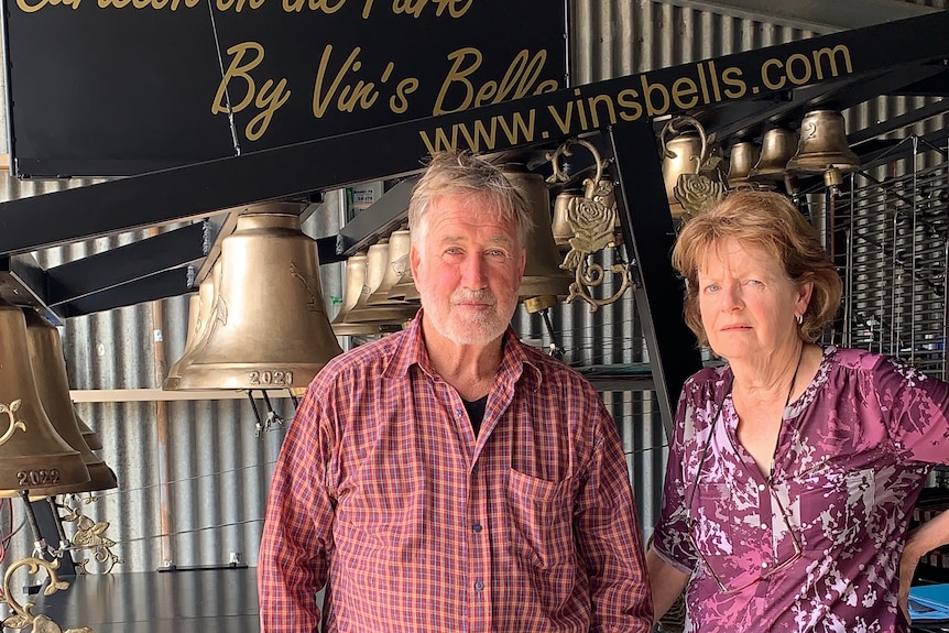 a man and woman stand in front of a black and gold bell instrument inside a workshed.