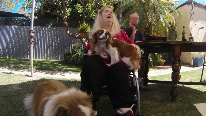 A woman sitting on a walking frame holding a small dog while a large dogs stands on grass