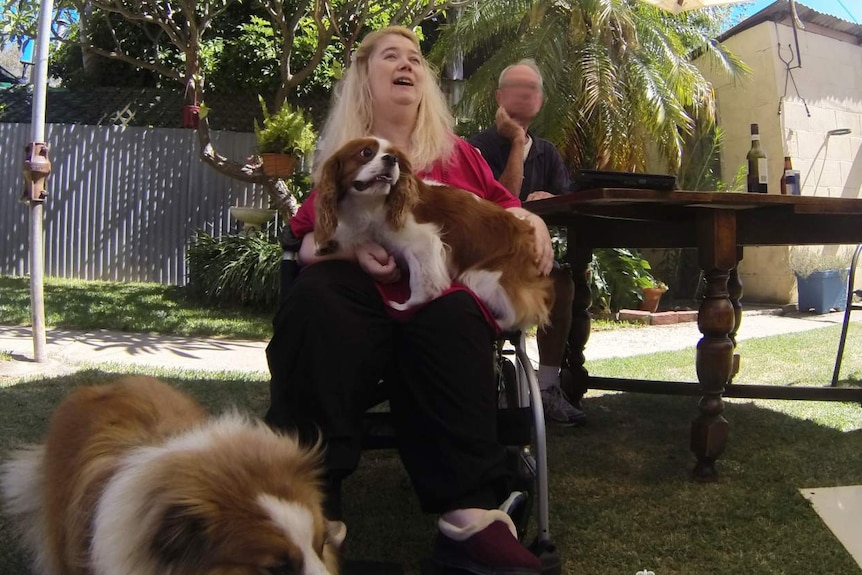 A woman sitting on a walking frame holding a small dog while a large dogs stands on grass