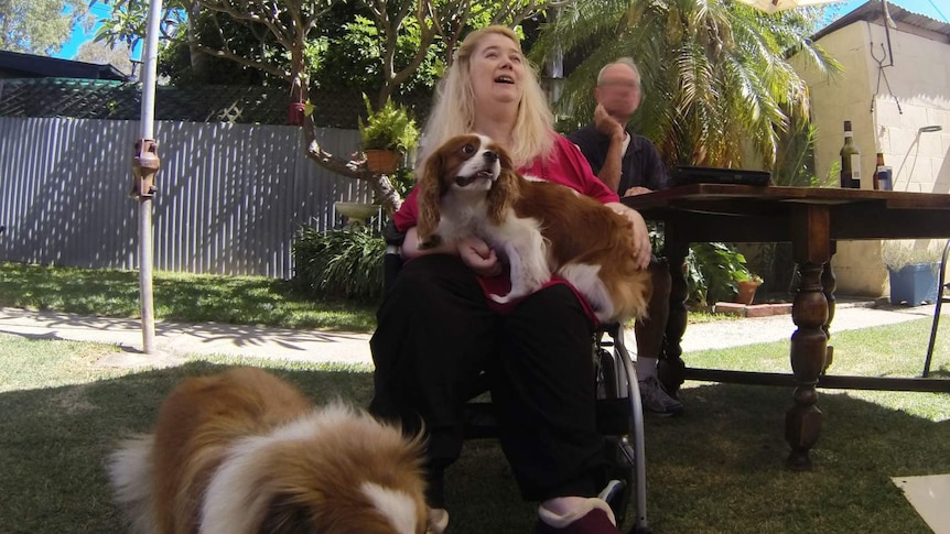 A woman sitting on a walking frame holding a small dog while a large dogs stands on grass