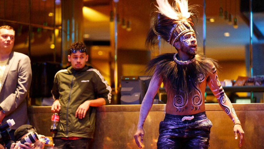 Interior of Hamer Hall, foyer. A man in black leather pants, bare torso and feathered headdress stands as if addressing crowd.