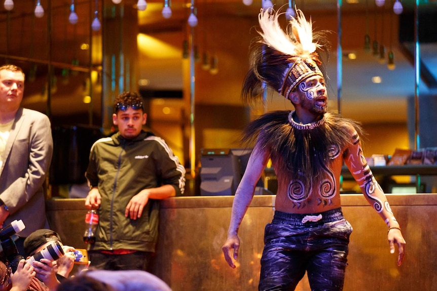 Interior of Hamer Hall, foyer. A man in black leather pants, bare torso and feathered headdress stands as if addressing crowd.