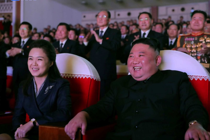 a still image from North Korean state TV showing Ri Sol Ju and Kim Jong Un seated in a theatre smiling while people behind stand