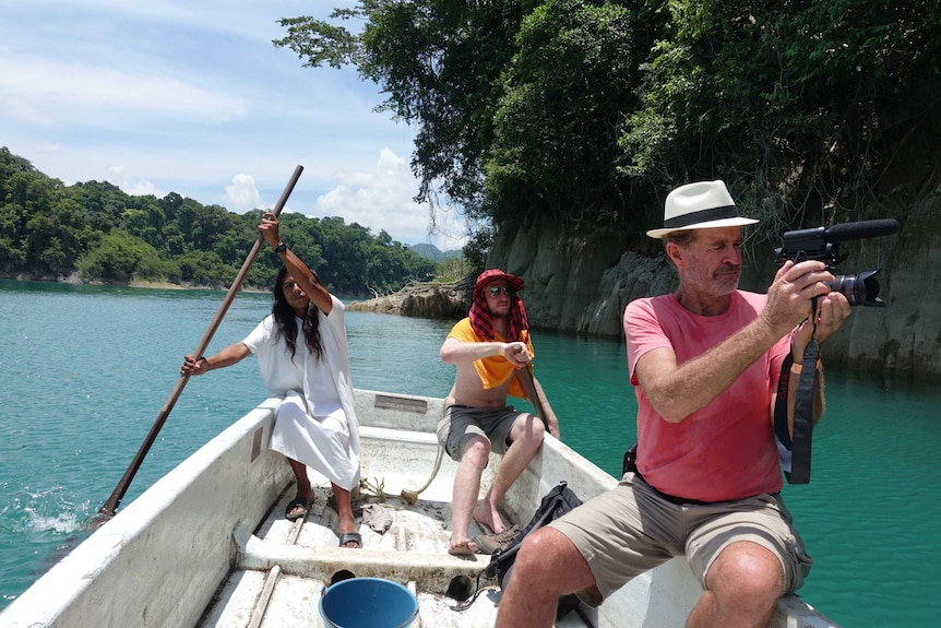 Three people on a small boat, as one man holds a video camera while on the water