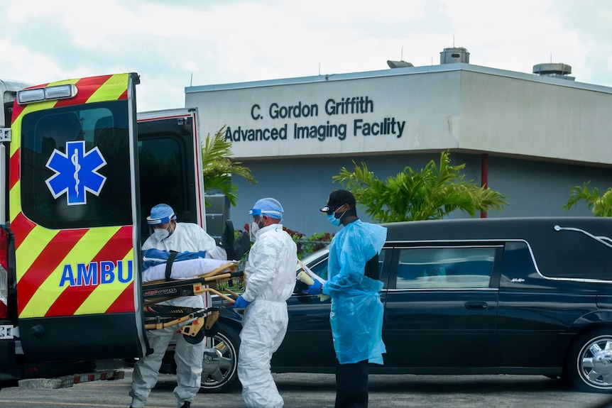 Medical staff take a patient on a stretcher out of an ambulance as a hearse pulls away.