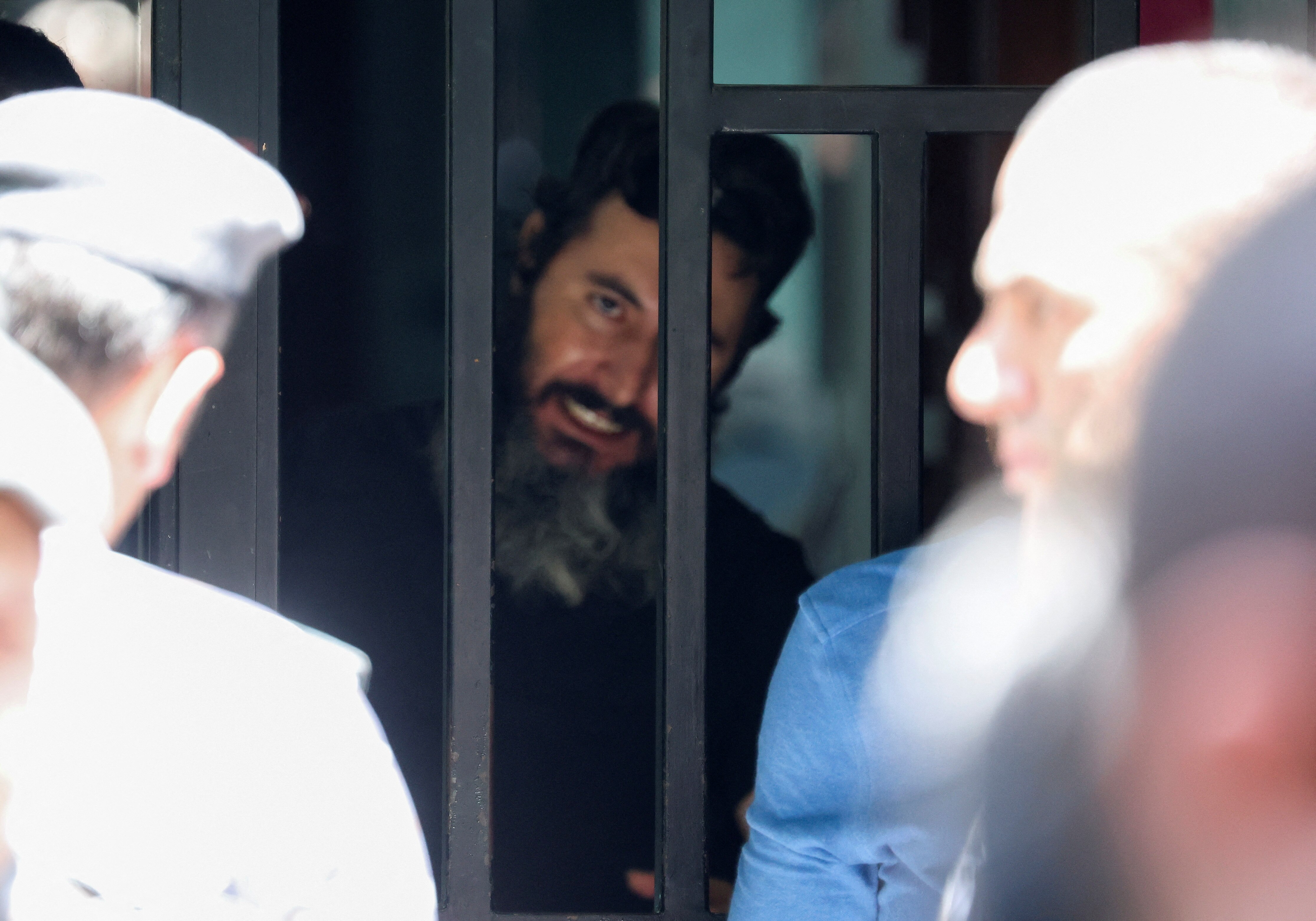 A man with black hair and a grey beard stares out intently through metal bars at crowd of people standing outside.