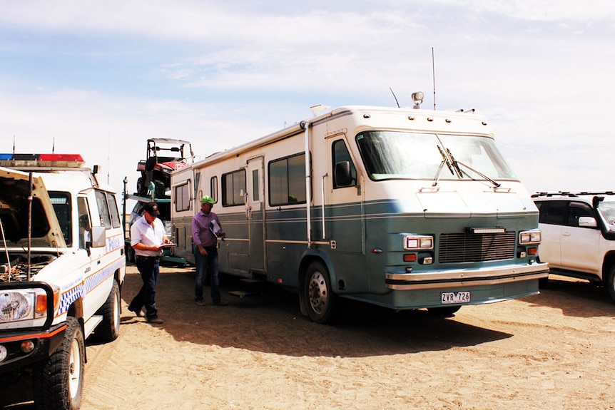 A large blue RV sitting in the red dirt of Bedourie, with a buggy and a car attached behind.