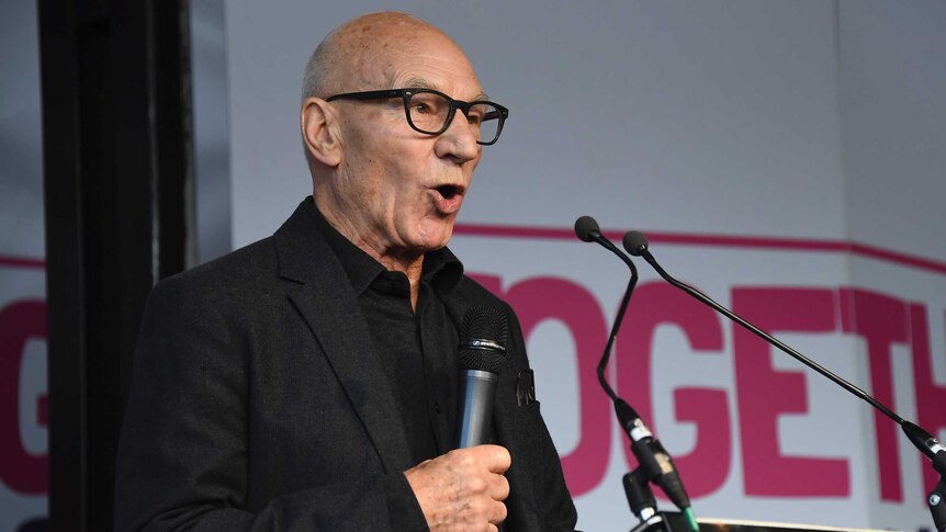Patrick Stewart stands at a lectern with a microphone in his hand. Behind him, a banner reads "together".