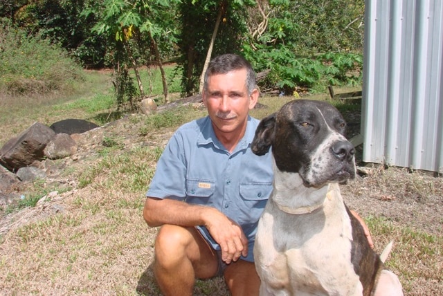 A man in a blue short sleeved shirt crouches beside a large brown and white dog in a garden.