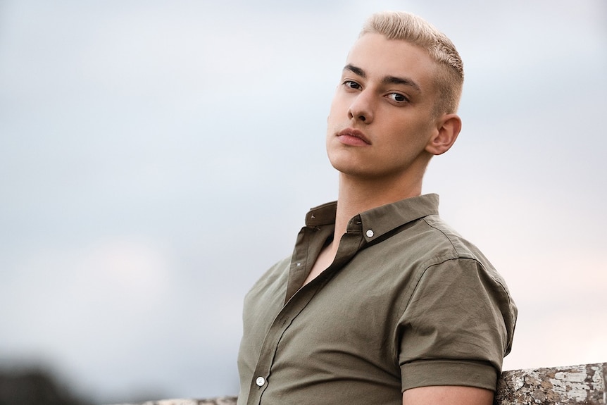 A young man with cropped blond hair stands in a paddock.