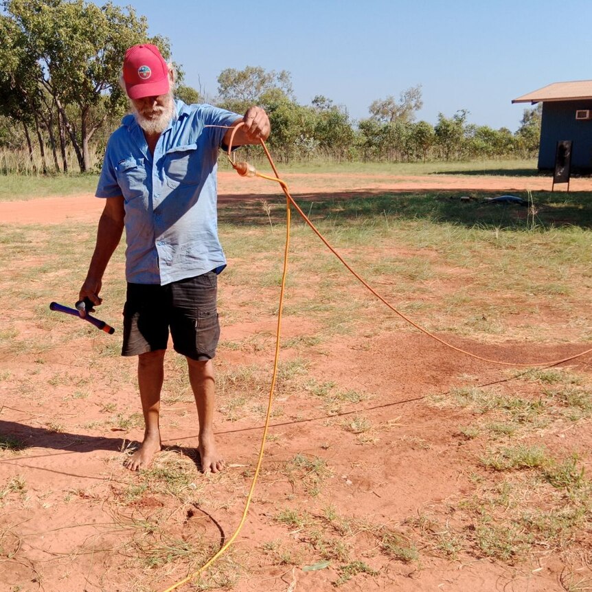A man in barefeet holds up a power cord running over red dirt 