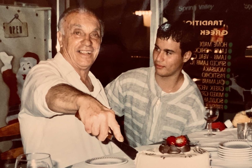An old photo of an older man sitting next to his adult grandson at a table in a restaurant with a cake on the table.