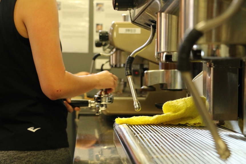 A young person, with face out of shot, presses ground coffee in front of two coffee machines.