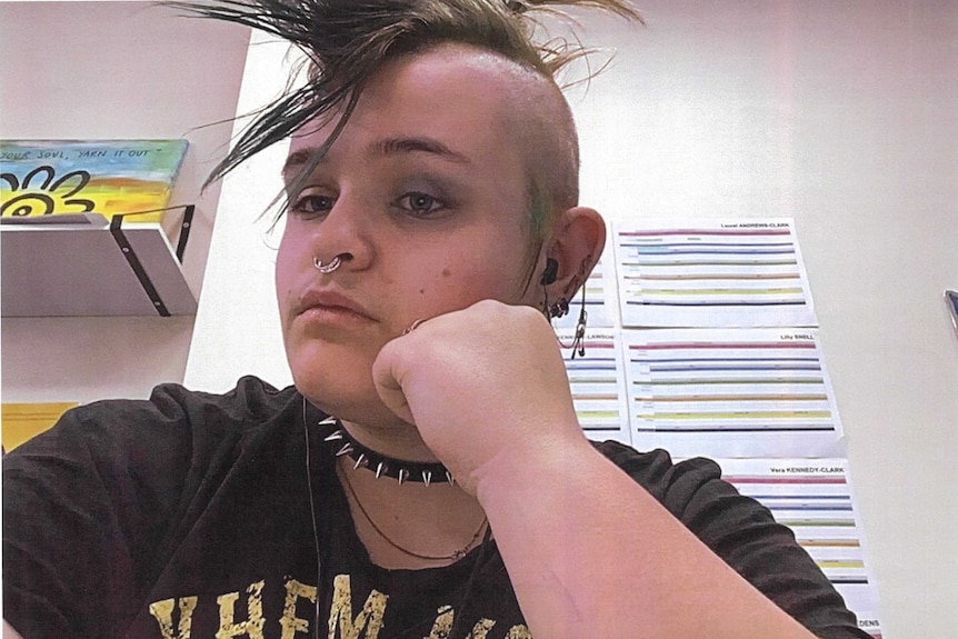 A boy wearing a black shirt and spiked collar rests his head on his hand. His hair is styled in a mohawk