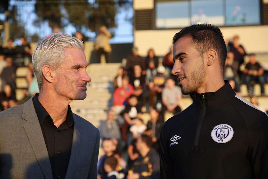A man with grey hair and a man in a black tracksuit smile and make eye contact on a soccer pitch.