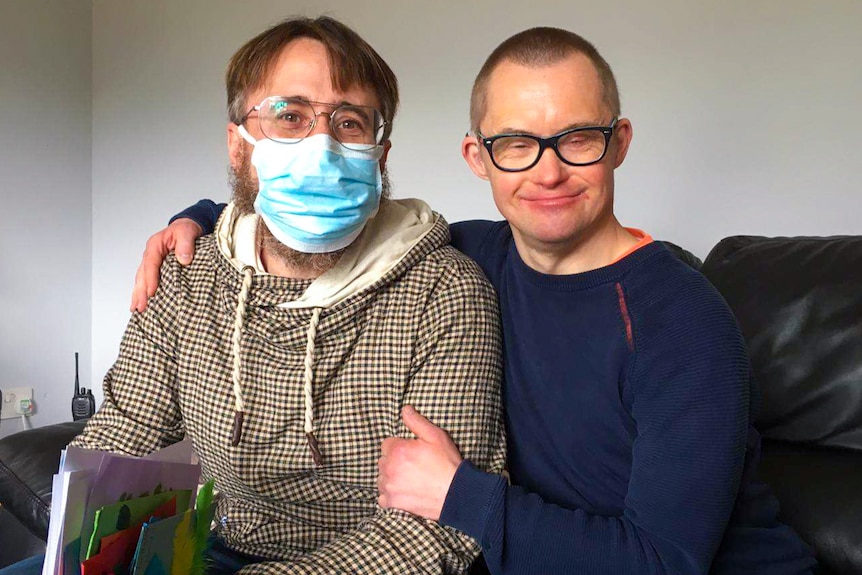 A man in a glasses and a mask sits with a man wearing glasses, who has his arm around him.