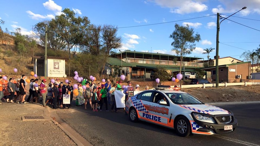 A police car leads a procession of people who are holding purple and orange balloons.
