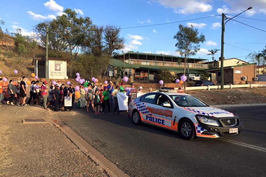 A police car leads a procession of people who are holding purple and orange balloons.