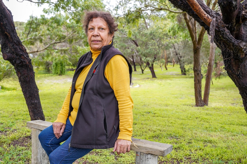 A woman sits on a wooden bench in a park, staring out over her left shoulder.