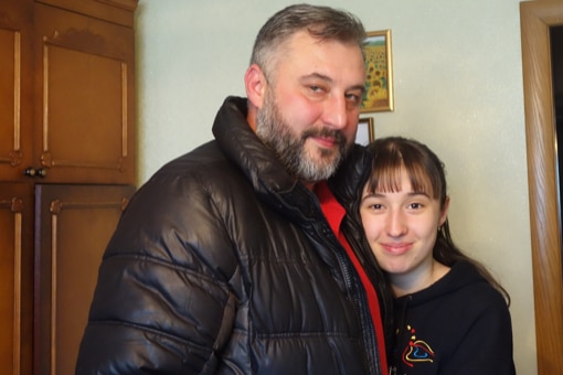 A man with close-cropped hair, bearded and wearing a leather jacket, hugging a teenage girl.