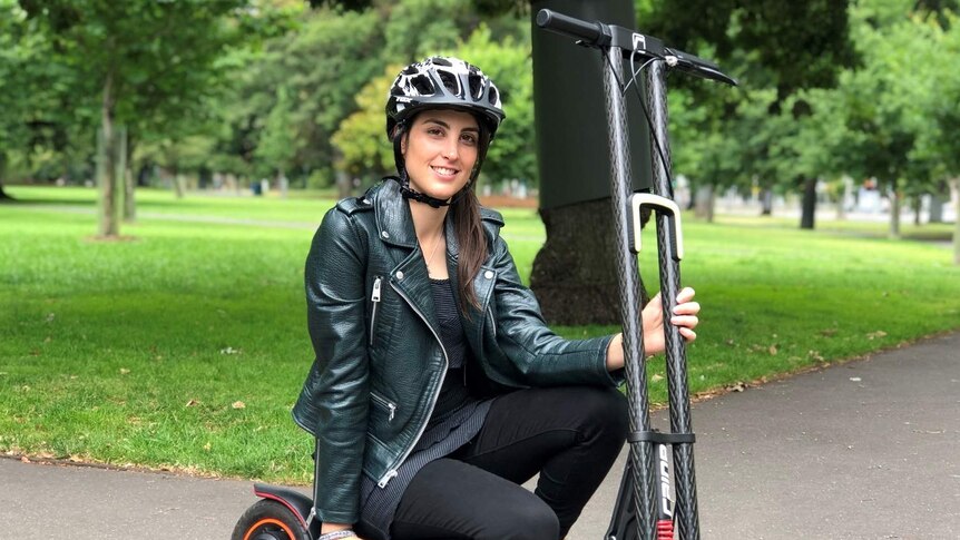 Michelle Mannering squats over the seat of an e-scooter wearing a helmet in a park and smiles at the camera.