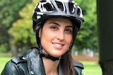Michelle Mannering squats over the seat of an e-scooter wearing a helmet in a park and smiles at the camera.