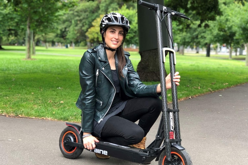 Michelle Mannering squats over the seat of an e-scooter wearing a helmet in a park and smiles at the camera.