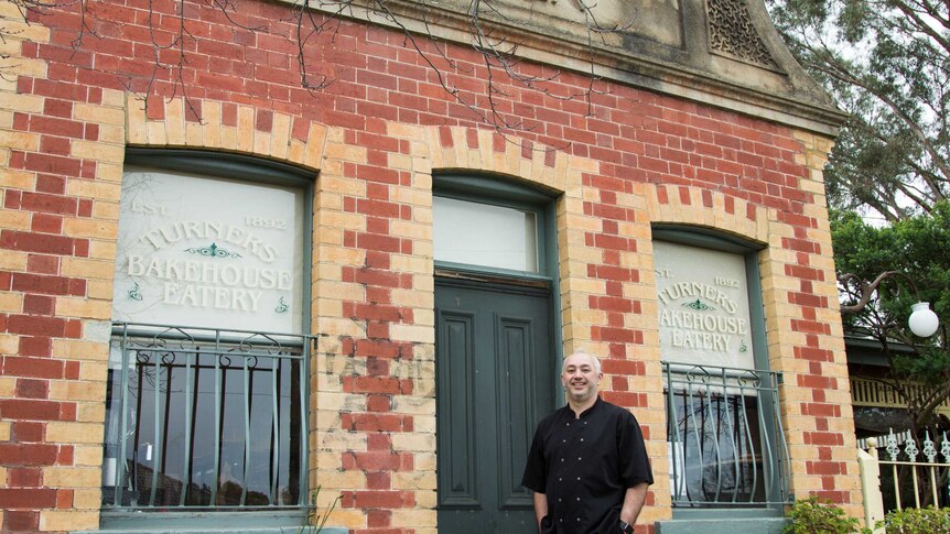 A man in chef's clothing stands outside a historic brick building, sign reads 'Turners Bakehouse Eatery'.