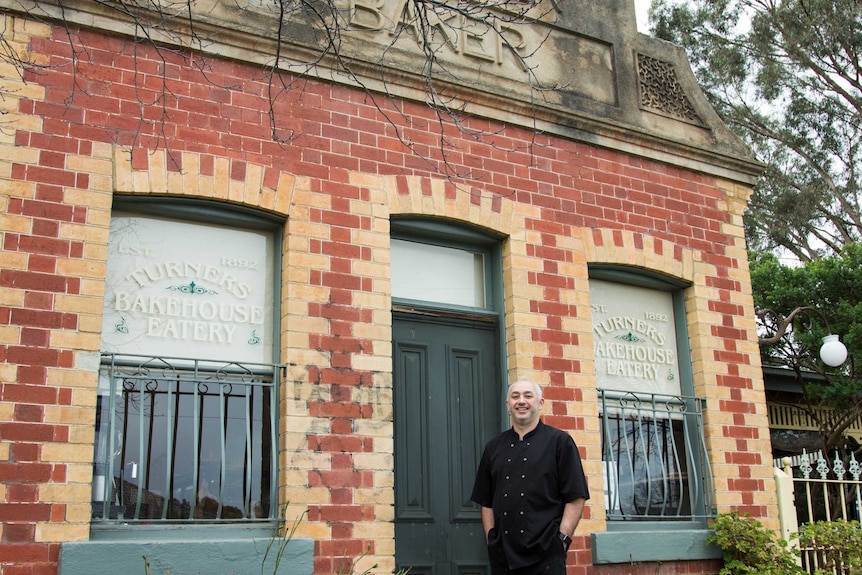 A man in chef's clothing stands outside a historic brick building, sign reads 'Turners Bakehouse Eatery'.