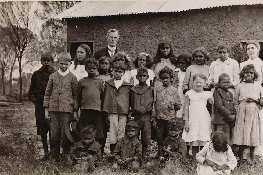 black and white photo of aboriginal children standing outside an old building with a white man and woman