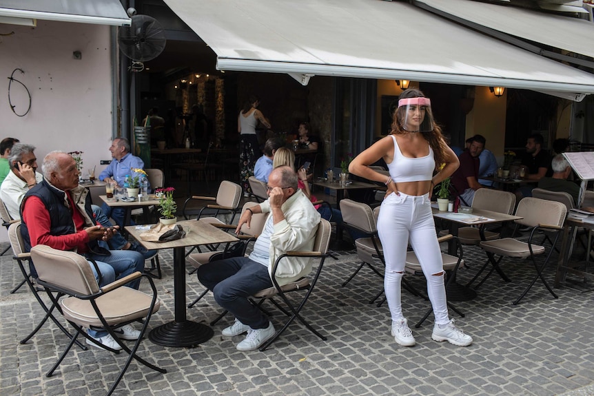 A young woman wearing a plastic face screen stands in front of an alfresco area filled with people drinking and talking.