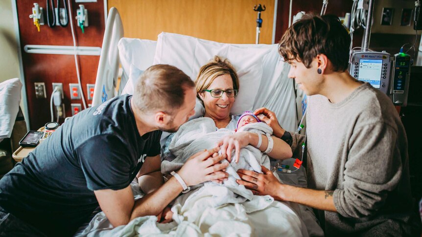 Two new dads flank woman in the middle holding baby on hospital bed.