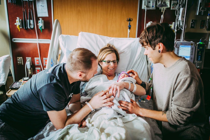 Two new dads flank woman in the middle holding baby on hospital bed.