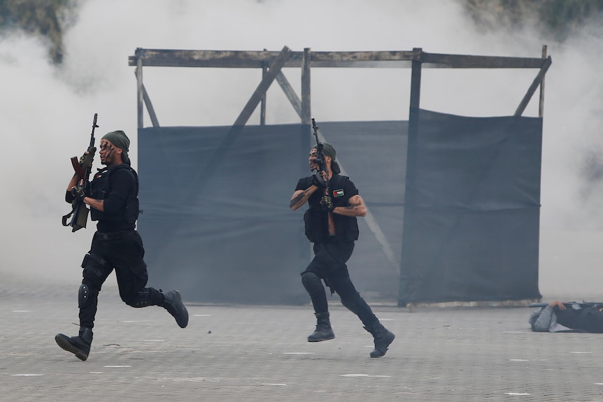 Two young men in black clothing carrying rifles run across a city square filled with smoke.