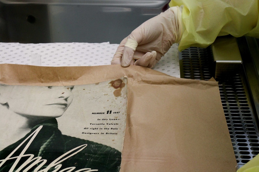 A gloved hand holds back brown paper beneath a book with a blood stain on the corner