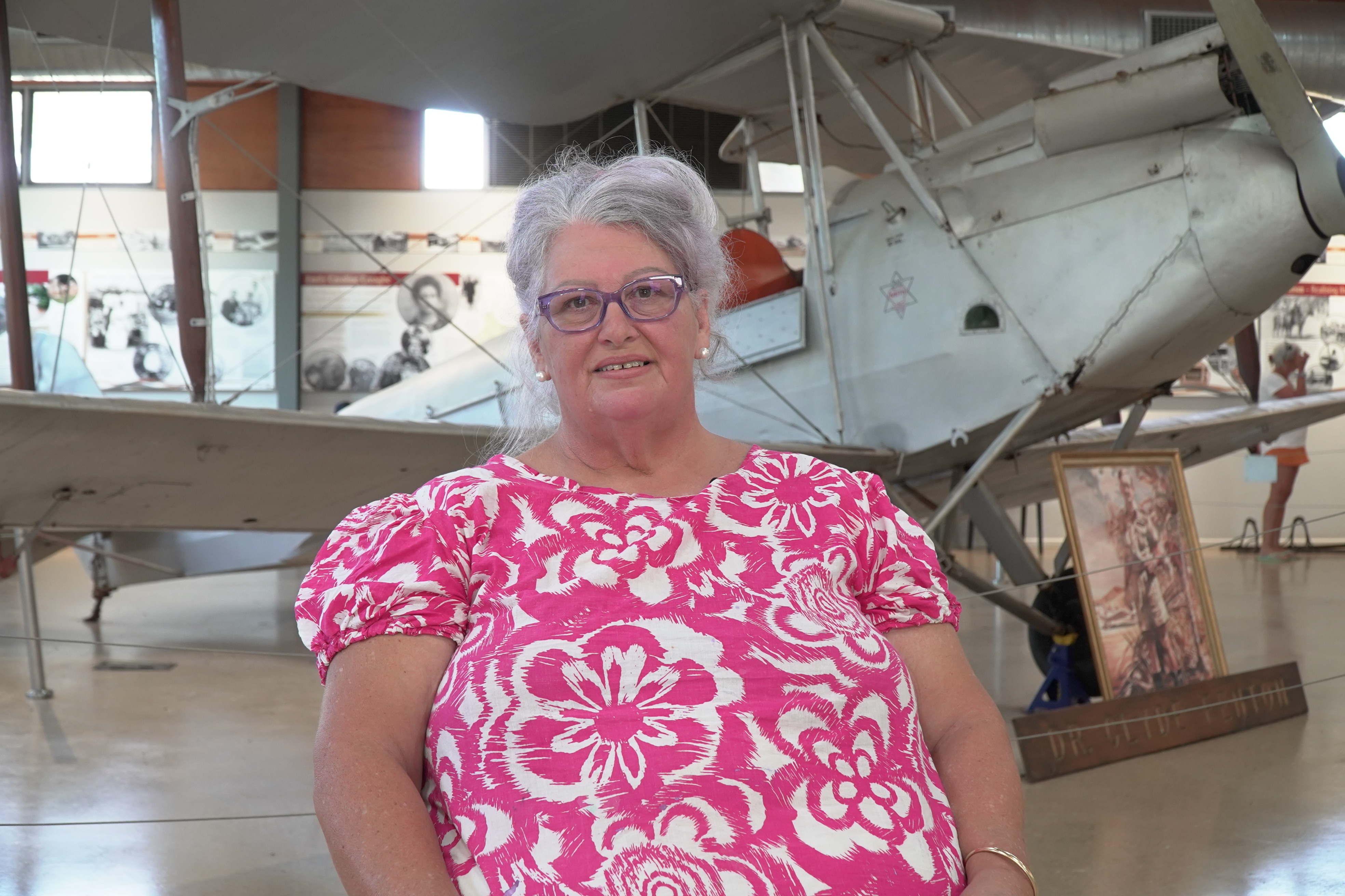 A woman in a pink and white patterned top sits in front of an old plane