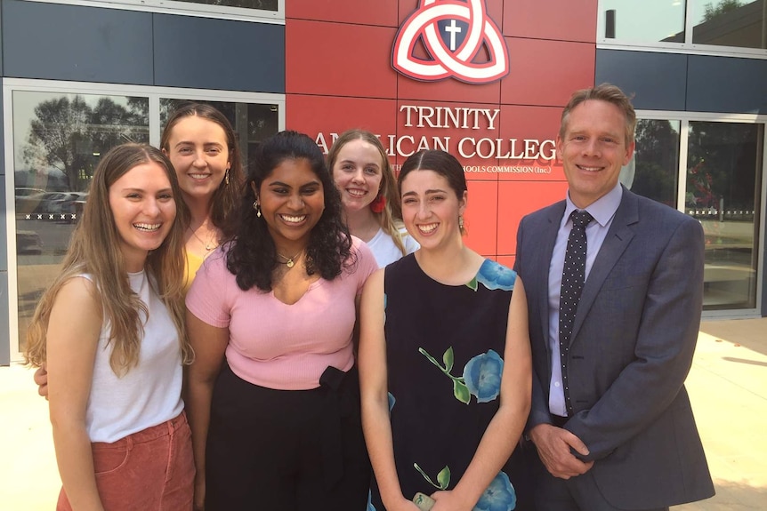 five girls stand next to man in suit in front of trinity anglican college sign