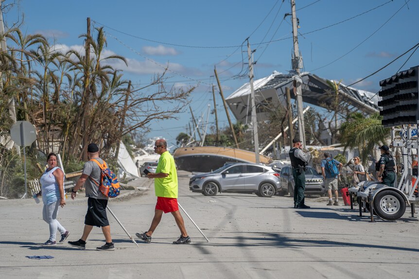 People walking across the street with trees in the background and hurricane damage.