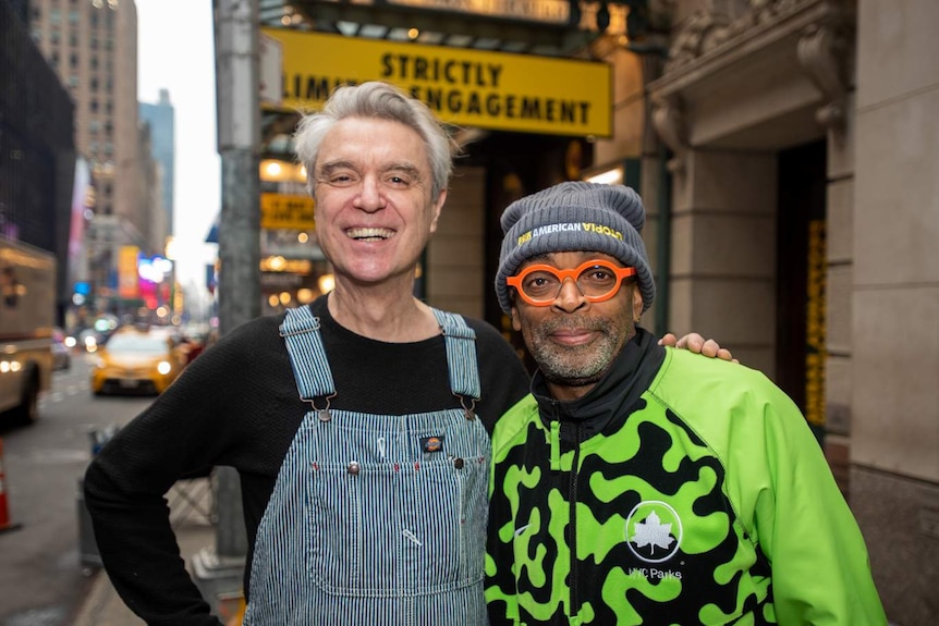 Outdoor street shot outside Broadway theatre with David Byrne in denim overalls and Spike Lee in grey beanie, both smiling.
