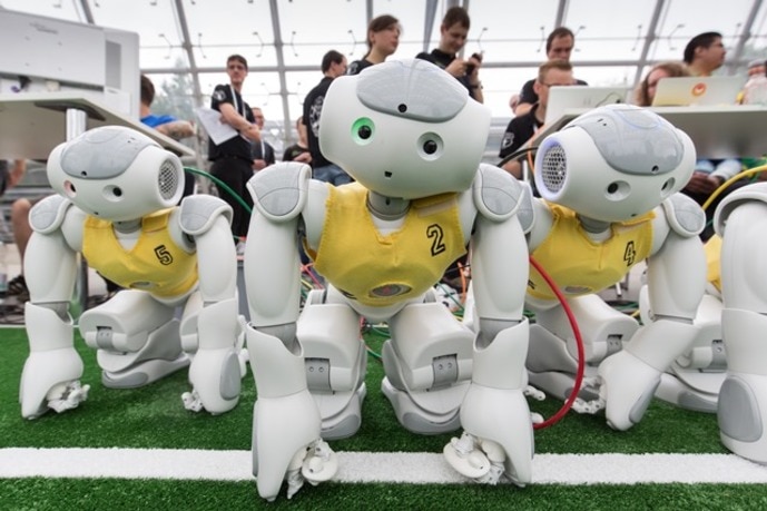 A robot football team wearing yellow vests waits on the side lines.