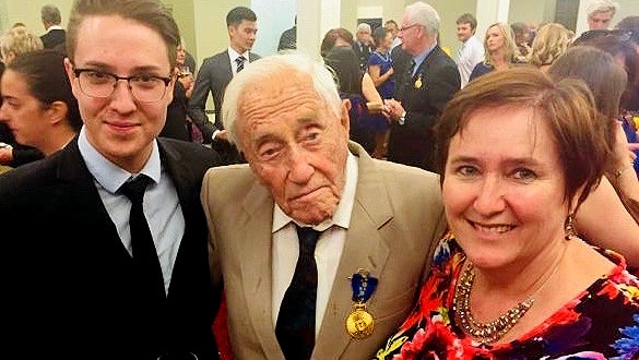 An elderly man in a tan suit with a medal on the breast standing next to a young man in a black suit and a woman in a flower top