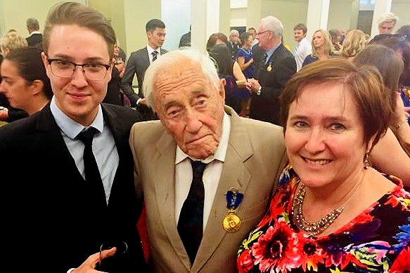 An elderly man in a tan suit with a medal on the breast standing next to a young man in a black suit and a woman in a flower top