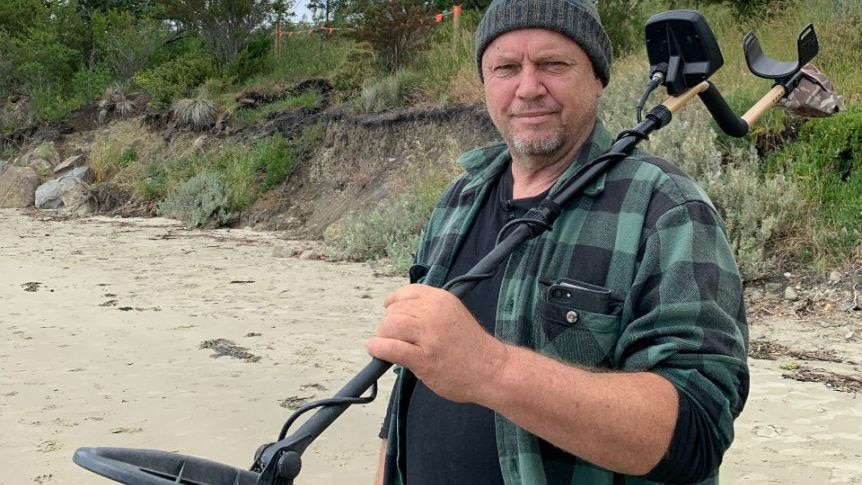 A man holds a metal detector over his shoulder on a beach