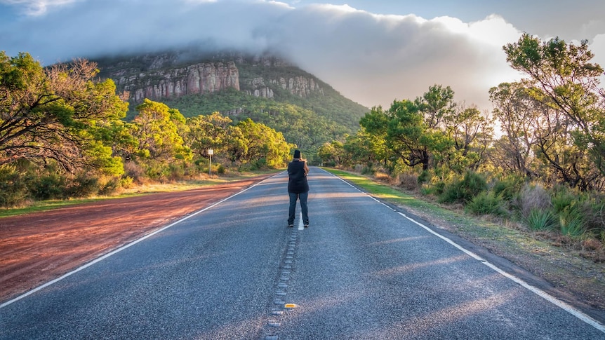 A man stands on a road at sunset. At the end of a road is a cloud-covered mountain.