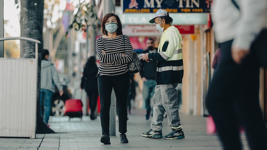A man and a woman wear masks while standing outside a shopping strip. 